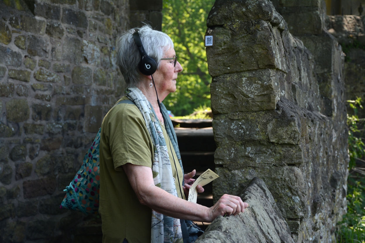 Lady next to an old stone wall, listening to audio on headphones, with her eyes closed.