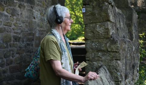 Lady next to an old stone wall, listening to audio on headphones, with her eyes closed.